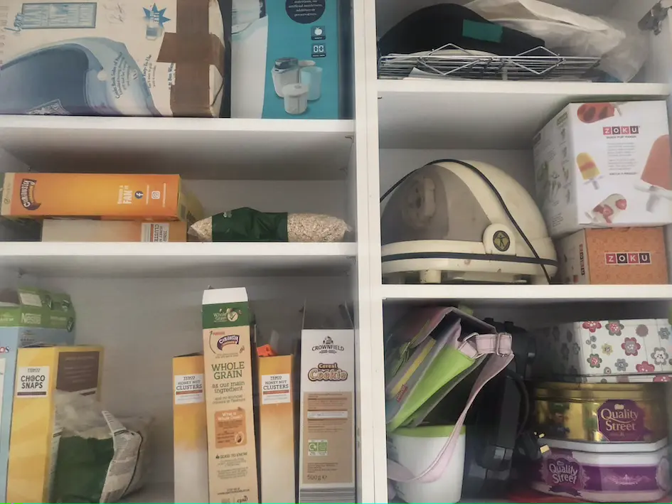 White kitchen cupboard with multiple shelves full of cereals, kitchen appliances and tins
